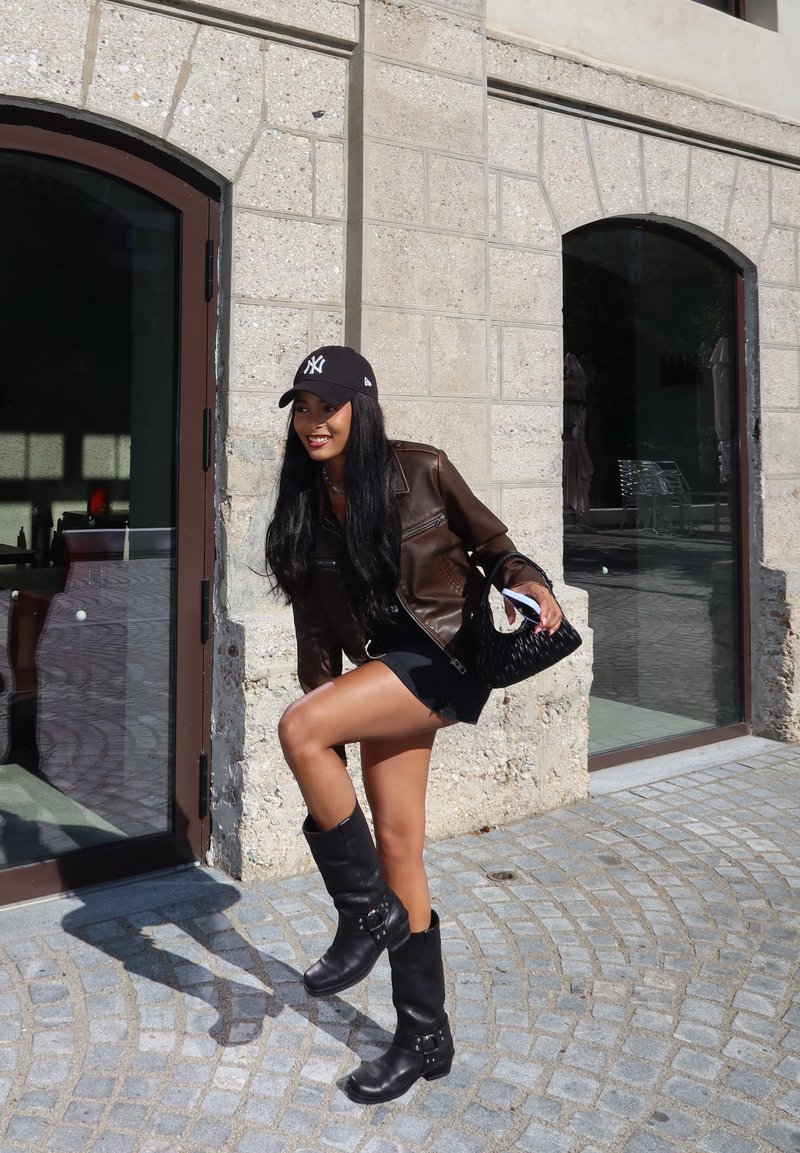 Καφέ δερμάτινο μπουφάν, μαύρο καπέλο με λογότυπο, μαύρη μίνι φούστα, μαύρες μπότες μέχρι το γόνατο με αγκράφες και μια πλεχτή μαύρη τσάντα. Φόντο από πέτρινο τοίχο.