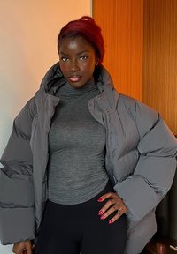 Femme aux cheveux roux portant un col roulé gris, une doudoune gris oversize, un pantalon noir et un vernis à ongles rouge, debout à l'intérieur.