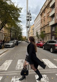 Žena s dlouhými vlasy v černém kabátu a botách přechází ulici přes přechod, drží černou kabelku, v pozadí vysoká věž a budovy.