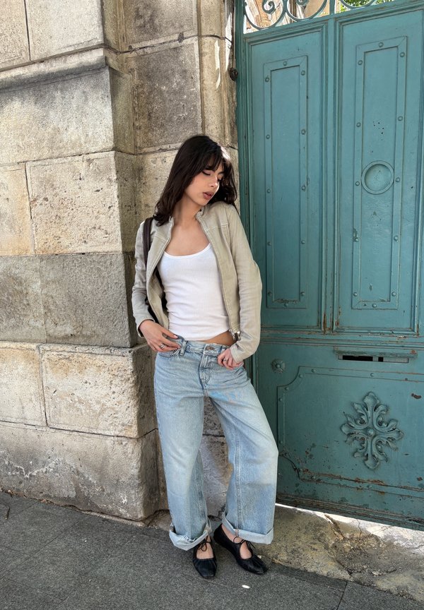 Jeune femme appuyée contre un mur en pierre près d'une porte turquoise ornée, portant une veste beige, un débardeur blanc, un jean ample bleu clair et des ballerines noires.
