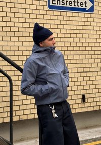 Veste zippée rayée bleu et blanc avec col haut, associée à un bonnet en tricot foncé et un pantalon noir. Porte-clés attaché à la ceinture.