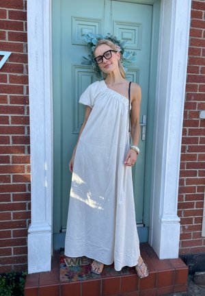 Mujer con gafas que lleva un vestido blanco de un solo hombro y sandalias, sonriendo frente a una puerta verde con una corona y una pared de ladrillo rojo.