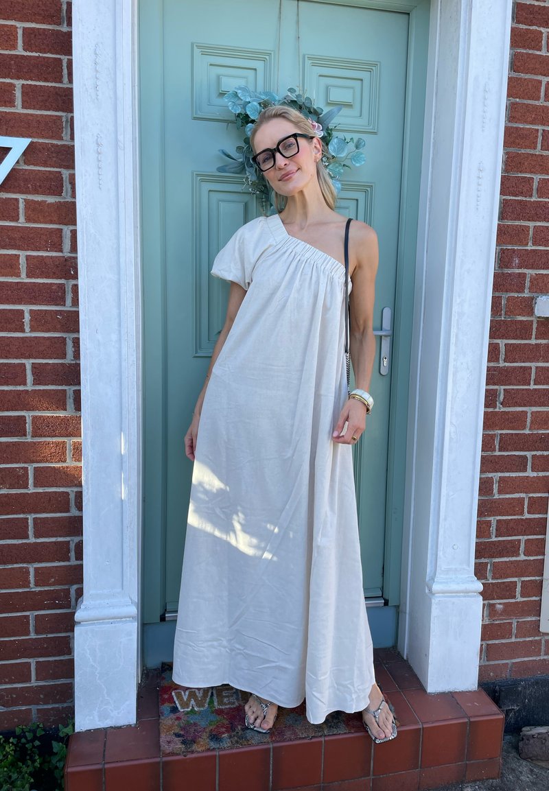 Donna con occhiali che indossa un abito bianco a spalla unica e sandali, sorride davanti a una porta verde con una ghirlanda e un muro di mattoni rossi.