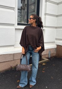 Donna con poncho marrone e occhiali da sole che tiene una borsa marrone, indossa jeans blu e scarpe marroni con la punta a punta, in piedi accanto a un edificio bianco.