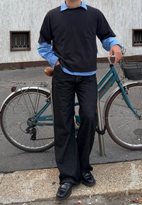 Maglietta nera a maniche corte oversize indossata sopra una camicia azzurra con colletto, abbinata a jeans scuri e scarpe nere. Sullo sfondo c'è una bicicletta turchese.