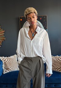 Jeune personne aux cheveux blonds bouclés portant une chemise blanche ample et un pantalon gris, debout devant un mur sombre avec des œuvres d'art et des coussins.