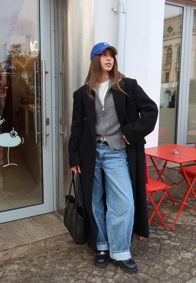 Mujer con gorra azul, abrigo largo negro, suéter gris con cremallera, vaqueros anchos, zapatos negros y bolso, de pie fuera de una cafetería con mesa y sillas rojas.
