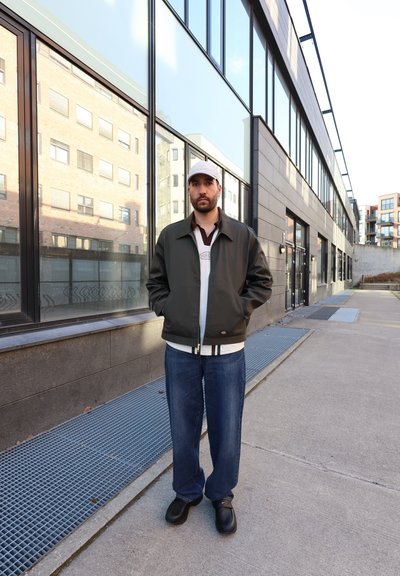 Hombre con chaqueta oscura, vaqueros azules y gorra clara que está de pie en una acera urbana junto a un edificio de vidrio bajo la luz del día.