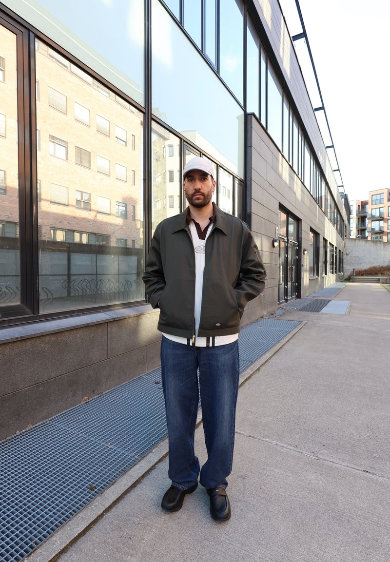 Homme portant une veste sombre, un jean bleu et une casquette claire, debout sur un trottoir urbain à côté d'un bâtiment en verre sous la lumière du jour.
