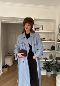 Femme portant un chapeau en fourrure marron et un manteau bleu clair, tenant un petit sac marron, debout dans une pièce moderne avec des étagères et des plantes.