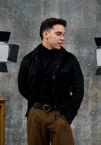 Homme en veste noire et col roulé avec un pantalon marron et une ceinture, debout contre un mur gris usé entre deux lampes de studio.