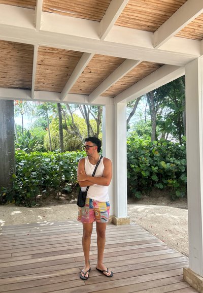 Una persona con una camiseta sin mangas blanca y pantalones cortos de patrón colorido está de pie sobre un suelo de madera en un área al aire libre luminosa con una vegetación exuberante.