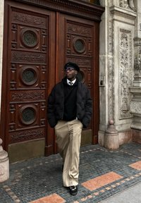 Sort pufferjakke over en sort sweater, beige vide bukser, sort beret og briller, stående foran udsmykkede trædøre.