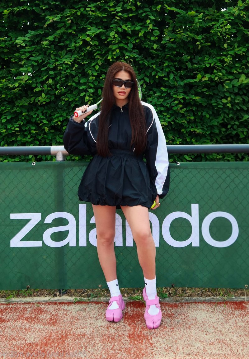 Jeune femme en veste de sport noire et short, tenant une raquette de tennis, debout sur un court en terre battue devant une clôture verte avec un panneau "Zalando".