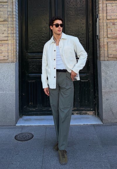 Hombre con chaqueta blanca, camisa blanca, pantalones grises y gafas de sol de pie frente a una puerta decorativa oscura en la acera de la ciudad.