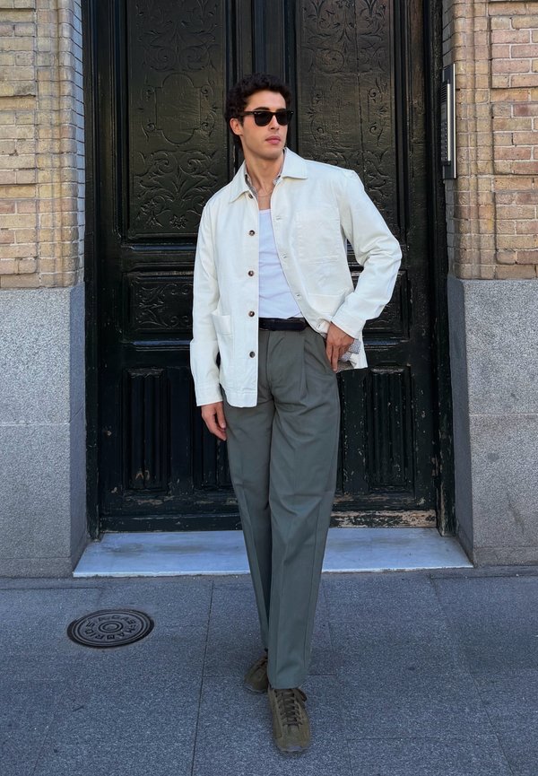 Homme portant une veste blanche, une chemise blanche, un pantalon gris et des lunettes de soleil debout devant une porte décorative sombre sur le trottoir de la ville.