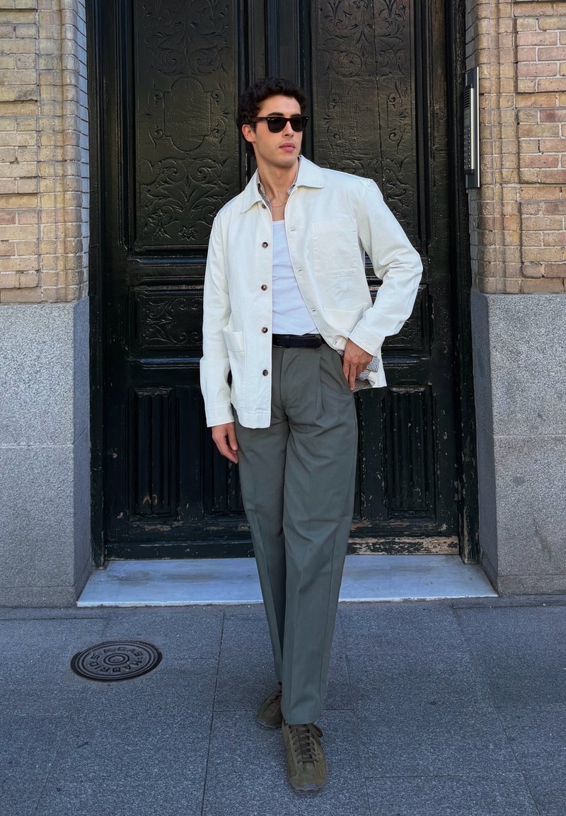 Homme portant une veste blanche, une chemise blanche, un pantalon gris et des lunettes de soleil, debout devant une porte décorative sombre sur le trottoir de la ville.