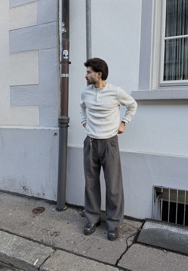 Man in white long-sleeve shirt and gray wide pants stands on sidewalk looking left beside gray building with window and drainpipe.