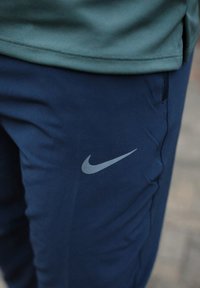 Gros plan d'une personne portant un pantalon Nike bleu marine avec le logo Swoosh visible près de la poche, associé à un haut vert.