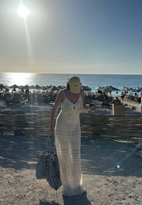 Długa, biała sukienka szydełkowa z dekoltem w kształcie łezki, uzupełniona żółtą czapką i torebką w niebieską kratkę. Tło plaży oświetlone słońcem.