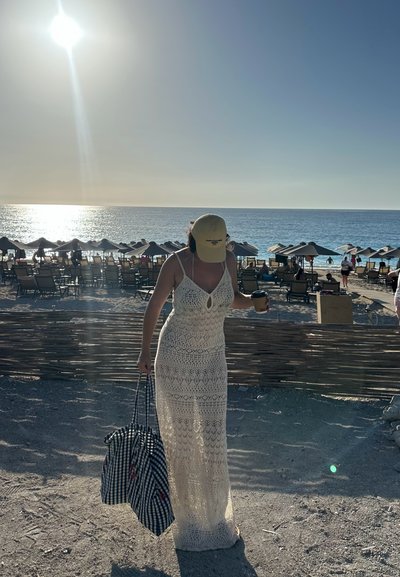 Largo vestido de ganchillo blanco con un diseño de ojal, complementado con una gorra amarilla y un bolso de cuadros azules. Fondo de playa iluminado por el sol.