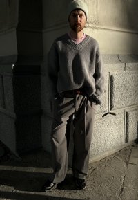 Pull gris côtelé, chemise rose en dessous, pantalon beige et baskets foncées. Debout contre un mur en pierre texturé dans une lumière douce.