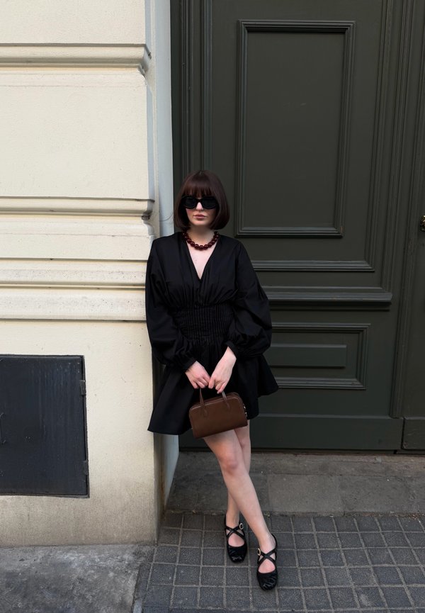 Mujer con vestido negro y gafas de sol está apoyada en la pared de un edificio, sosteniendo un bolso marrón, con zapatos negros y un collar rojo de cuentas.