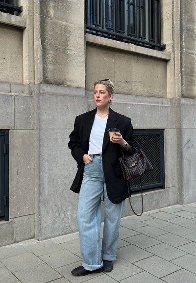 Mujer con cabello rubio corto, vestida con un blazer negro oversize y jeans azul claro, sosteniendo una taza de café y un bolso marrón tejido en la acera de la ciudad.