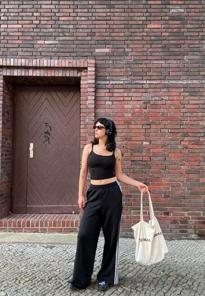 Top corto negro y pantalones de chándal anchos negros con rayas blancas. Sosteniendo una bolsa de tela beis. El fondo presenta una puerta con patrón de chevron marrón.