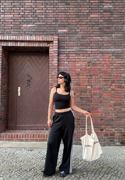 Top corto negro y pantalones de chándal anchos negros con rayas blancas. Sosteniendo una bolsa de tela beis. El fondo presenta una puerta con patrón de chevron marrón.