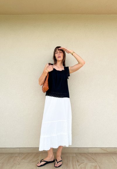 Top negro con encaje y falda blanca de longitud hasta el tobillo con diseño en capas. Un bolso de mano tejido marrón y unas chanclas negras completan el look.