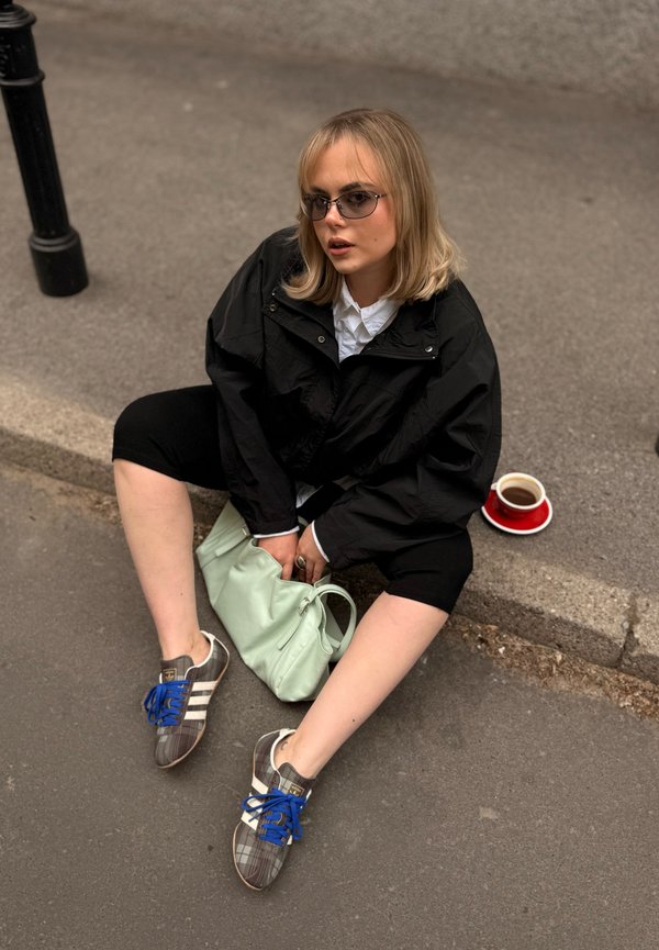 Giovane donna con occhiali da sole, giacca nera e sneakers seduta sul bordo del marciapiede che tiene una borsa verde menta con una tazza di caffè sul marciapiede accanto a lei.