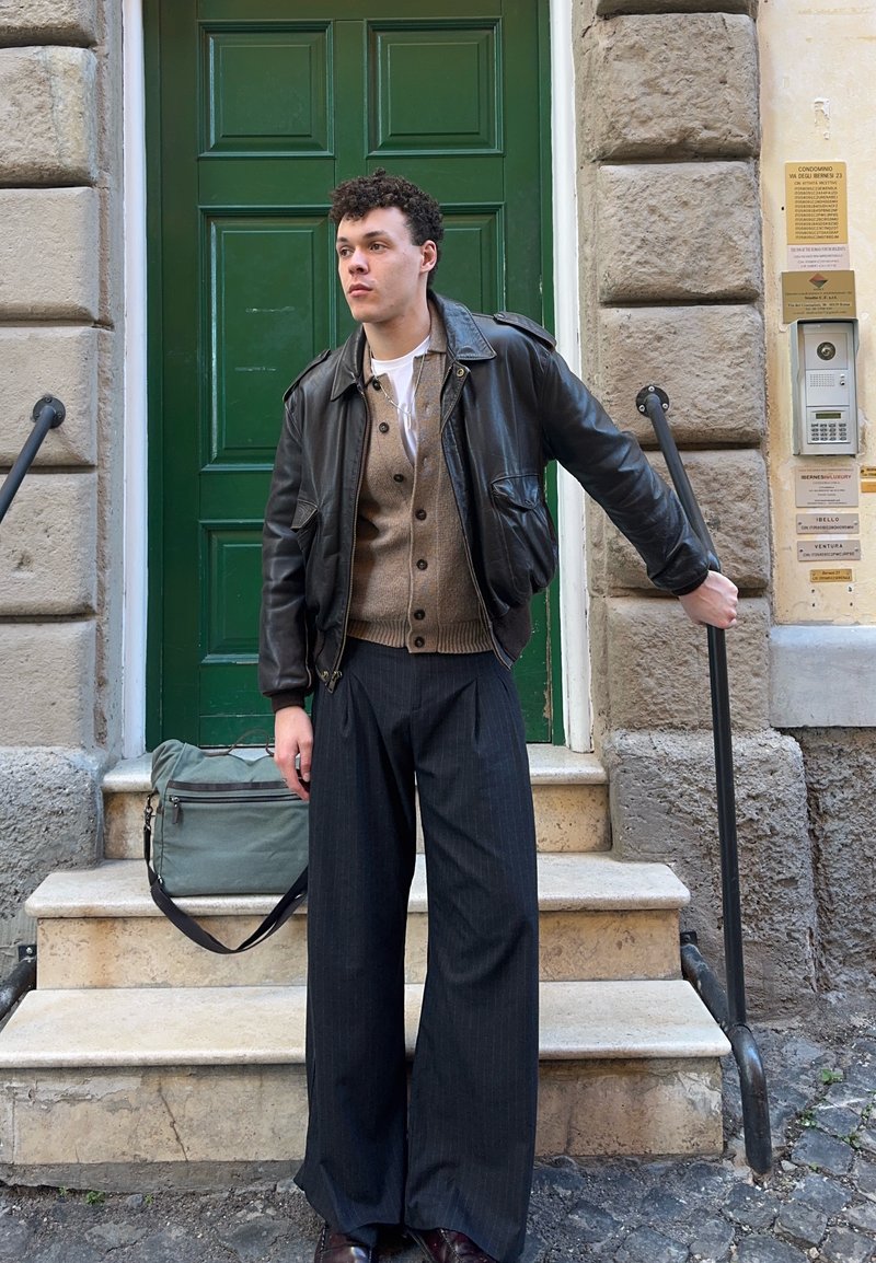 Jeune homme en veste en cuir noire, cardigan beige et pantalon à jambes larges, debout sur des marches en pierre, tenant la rampe en métal à côté d'une porte verte.