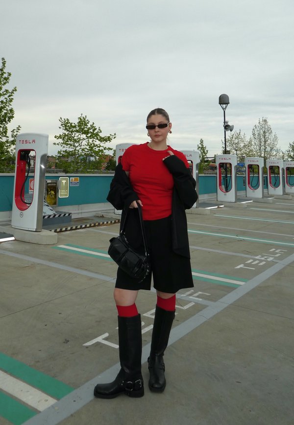 T-shirt rouge ajustée, veste noire surdimensionnée, shorts noirs, bottes noires montantes avec boucles, chaussettes rouges, sac à main noir. Stations de recharge en arrière-plan.