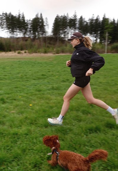 Corredor con chaqueta negra y pantalones cortos, calcetines blancos y zapatos gris claro, en movimiento sobre un césped verde con un perro marrón cerca.