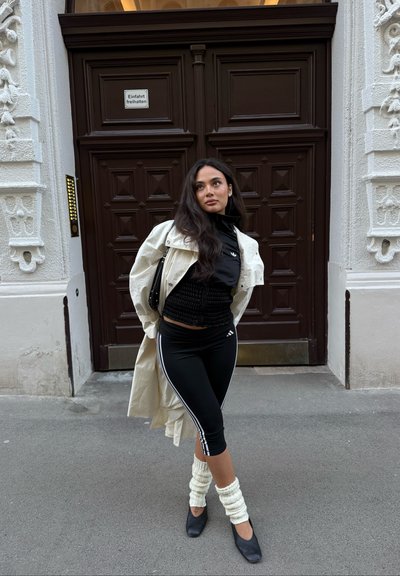 Mujer con ropa deportiva negra, abrigo largo blanco y calentadores de piernas, de pie frente a unas elaboradas puertas dobles de madera en la acera de la ciudad.