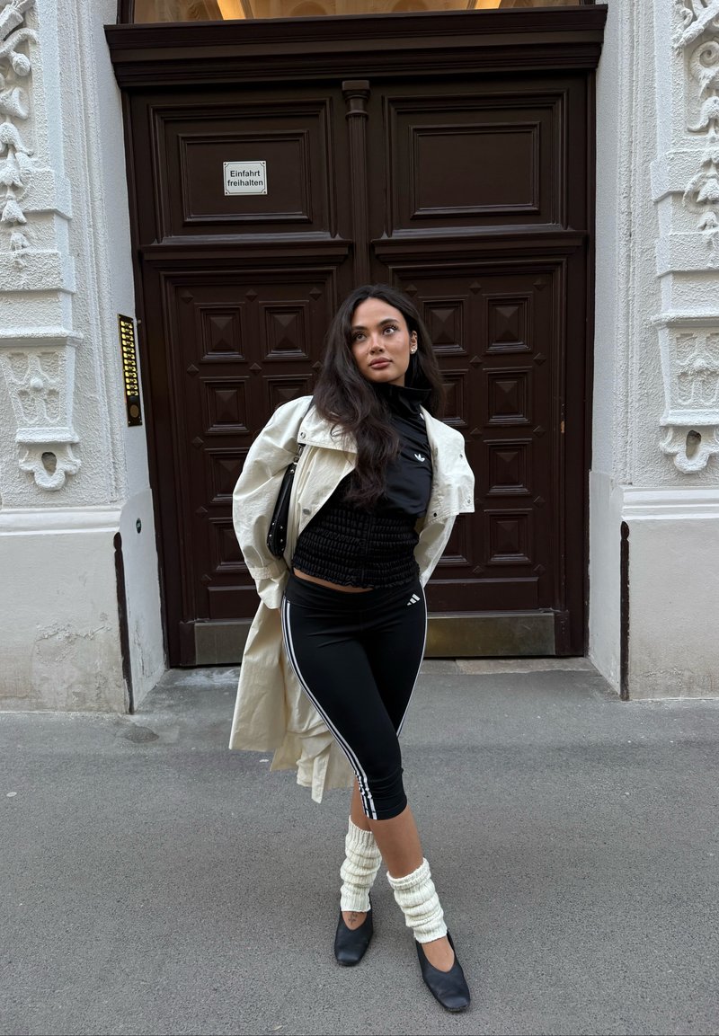 Femme vêtue de noir sportif, long manteau blanc et guêtres, se tient devant des portes doubles en bois ornées sur le trottoir de la ville.