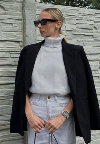 Blazer preto sobre um pulôver canelado cinza claro, calças brancas de cintura alta e óculos de sol retangulares pretos; mãos segurando a alça de uma bolsa.