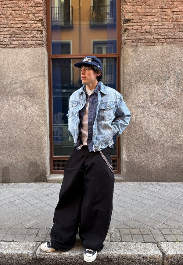 Un tânăr care poartă o jachetă din denim, șapcă, cămașă în carouri și pantaloni negri largi stă pe marginea trotuarului în fața unei clădiri din cărămidă și sticlă.