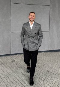 Szary dwurzędowy żakiet z czarnymi guzikami, zestawiony z czarnymi spodniami i czarnymi butami, idący po texturedkamiennej nawierzchni.