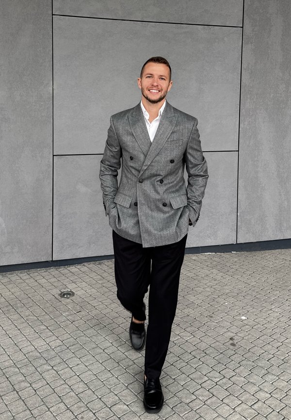 Siv dvojni blazer s črnimi gumbi, usklajen s črnimi hlačami in črnimi čevlji, hodi po teksturirani kamniti površini.