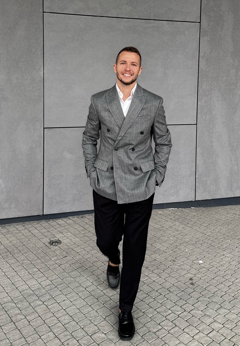 Blazer cinza de dois botões com botões pretos, combinado com calças pretas e sapatos pretos, a caminhar sobre uma superfície de pedra texturizada.