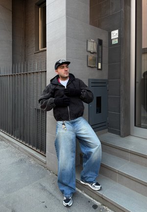 Hombre con chaqueta negra, guantes y gorra apoyado en la esquina de un edificio de azulejos grises, cerca de escaleras y una puerta de metal.