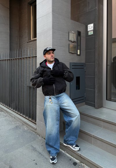 Hombre con chaqueta negra, guantes y gorra apoyado en la esquina de un edificio de azulejos grises, cerca de escaleras y una puerta de metal.