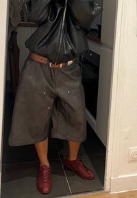 Hnedé kožené šortky s výrazným prešívaním a dvoma prednými vreckami, doplnené červenými teniskami. Pozadie zobrazuje kuchynské prostredie.