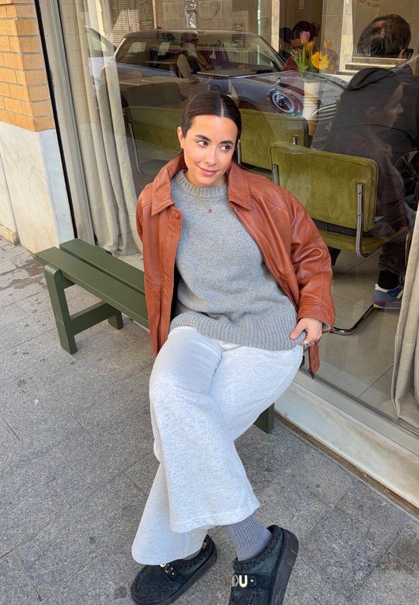 Γυναίκα με καφέ δερμάτινο μπουφάν και γκρι πουλόβερ κάθεται σε πράσινο παγκάκι έξω από καφέ με γυάλινες βιτρίνες που αντανακλούν ανθρώπους και αυτοκίνητο.