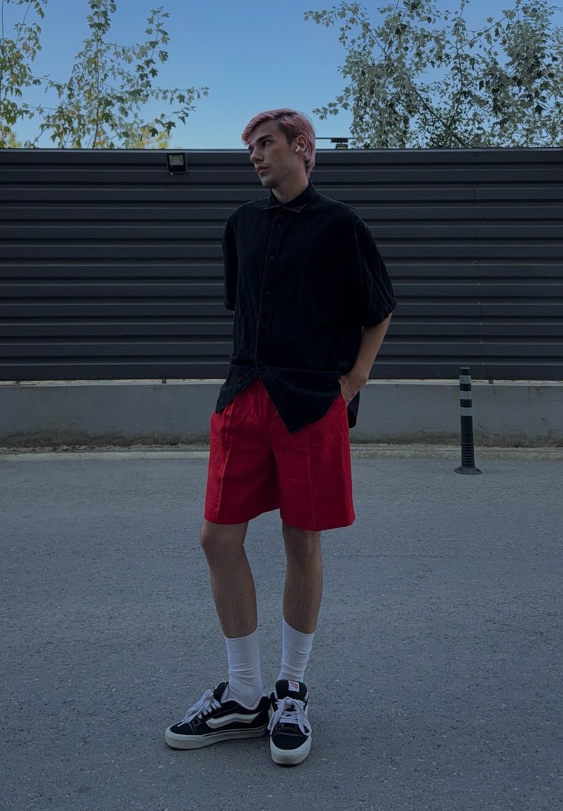 Jeune homme aux cheveux roses portant une chemise noire, un short rouge, des chaussettes blanches et des baskets noires, debout à l'extérieur sur un trottoir gris.