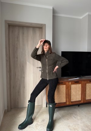 Mujer vistiendo chaqueta verde oscuro, pantalones negros, botas de lluvia verdes y gorra, posando en interiores cerca de un armario de madera y una puerta cerrada.
