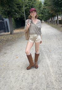 Des bottes marron arrivant au genou, un short beige avec des dentelles, et un pull léger gris clair à boutons. Un sac fourre-tout marron et une casquette bordeaux complètent le look.