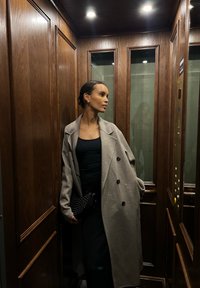 Sobretudo cinza com botões grandes, usado sobre uma blusa preta e saia preta. Clutch preta com tachas. Interior de elevador em madeira com painéis de vidro.